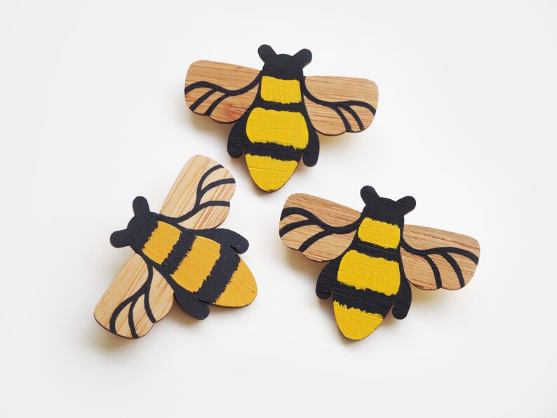 May include: Three wooden bee ornaments painted with black and yellow stripes. The bees have wings and are made of natural wood.