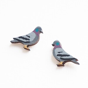 May include: Two wooden pigeon-shaped earrings. The earrings are painted gray with black wing details, a purple and blue head, and tan feet. The earrings are on a white background.