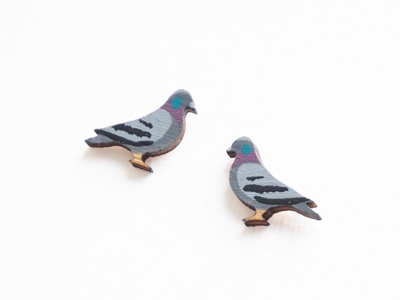 May include: Two wooden pigeon-shaped earrings. The earrings are painted gray with black wing details, a purple and blue head, and tan feet. The earrings are on a white background.