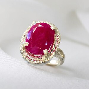 May include: A silver ring with a large, oval ruby gemstone. The ruby is a deep red hue, encircled by smaller, clear gemstones. The ring's band has a textured design, set against a white background.