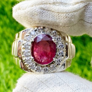 May include: A silver ring with a large, oval ruby gemstone encircled by smaller, clear gemstones. The ring has a ribbed design on the sides. The ring is held against a white background.