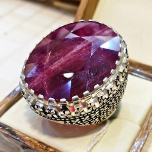 May include: A silver ring featuring a large, faceted, deep red ruby gemstone. The ring has a detailed, intricate design around the stone.