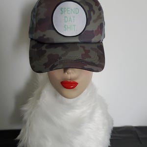 May include: A camouflage print trucker hat featuring a circular white patch on the front panel. The patch contains the green text $PEND DAT $HIT. The hat has a structured front, a curved brim, and a mesh back panel for ventilation and breathability.