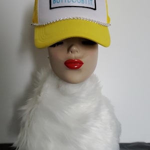 May include: A bright yellow and white trucker hat featuring a rectangular patch with the text I MAY BE WRONG BUT I DOUBT IT in light blue letters. The hat includes a silver chain trim along the base of the crown and a yellow curved brim.