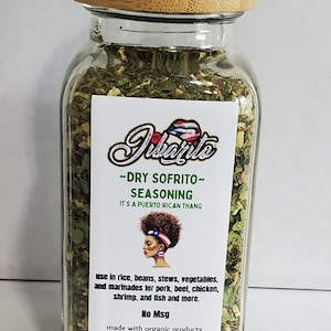 May include: A clear glass jar filled with dry sofrito seasoning, topped with a wooden lid. The label features the text "Dibants - DRY SOFRITO - SEASONING" and a graphic of a woman with a Puerto Rican flag in her hair. The jar contains 6.0 oz (223 g) of seasoning.