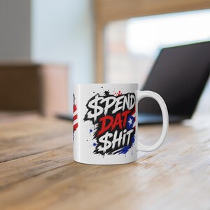 May include: A white ceramic mug featuring a graphic design with the text "$PEND DAT $HIT" in bold, distressed black and red lettering. The design includes paint splatter accents and a partial flag motif with a white star on a blue background.