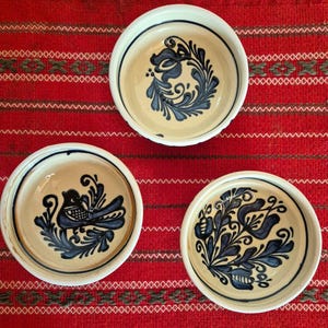 Puede incluir: Tres cuencos de cerámica con interior color crema y borde blanco. Cada cuenco presenta un diseño floral o de pájaro azul diferente. Los cuencos están dispuestos sobre un textil estampado en rojo y blanco.