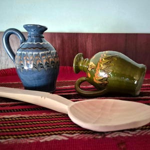 May include: A blue ceramic pitcher with a handle and decorative patterns, next to a green ceramic pitcher and a large wooden spoon. The items are on a red and black patterned textile, with a light blue and brown background.