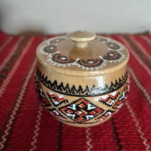 Peut inclure: Un petit récipient rond en bois avec un couvercle. Le récipient est peint avec un motif géométrique rouge, noir et blanc. Le couvercle a un petit bouton sur le dessus. Le récipient est sur une surface rayée rouge et blanche.