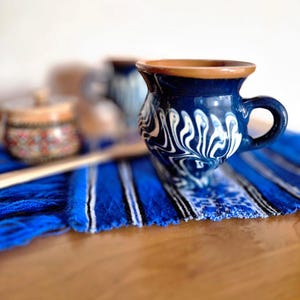 May include: A blue ceramic mug with a white swirl design and a brown rim rests on a blue and white striped textile. A small, decorated container and a wooden spoon are in the background. The mug has a handle.