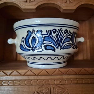 May include: Cream-colored ceramic bowl with blue floral and leaf designs. The bowl has two small handles and is set on a wooden shelf. The blue designs are hand-painted, adding a unique touch to the piece.