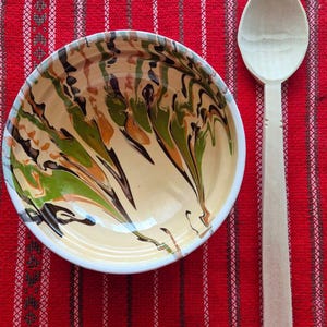 May include: A round ceramic bowl featuring a marbled pattern of cream, olive green, orange, and dark brown swirls, accompanied by a plain, light-coloured wooden spoon with a smooth, unfinished texture and a rounded handle and bowl shape.