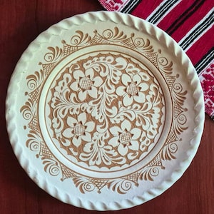 Pode incluir: Um prato redondo de cerâmica com superfície creme, gravuras florais castanhas detalhadas e uma borda com textura de corda. O design central apresenta flores e padrões de folhas em espiral como uma peça decorativa.