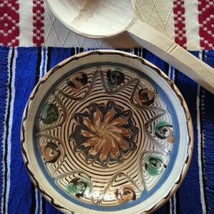 Peut inclure: Un bol en céramique rond présentant un motif floral central dans des tons de brun, bleu et vert, entouré de lignes concentriques et d'un bord festonné, accompagné d'une cuillère en bois clair à la finition lisse et sculptée.