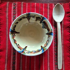 May include: A cream-coloured ceramic bowl with a colourful rim design, alongside a wooden spoon. The bowl and spoon are placed on a red and white striped placemat, creating a rustic, handcrafted aesthetic.