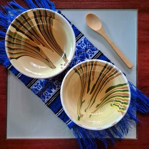 May include: Two ceramic bowls with cream interiors and a decorative pattern of brown, green, and tan lines. They sit on a blue and white patterned textile with blue fringe, next to a wooden spoon.