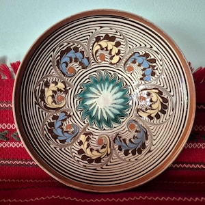 May include: A decorative ceramic plate with a brown rim and intricate patterns. The plate features a central floral design in teal and white, surrounded by swirling black and white lines and stylized floral motifs in yellow and blue.