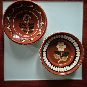 Puede incluir: Dos cuencos redondos de terracota con bordes festoneados. El cuenco superior presenta un motivo floral central en blanco, amarillo y verde. El cuenco inferior muestra una flor blanca con centro amarillo, rodeada por un borde de formas ovaladas blancas.