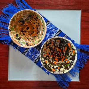 May include: Two handcrafted ceramic bowls with colourful, abstract designs. The bowls are placed on a blue and white patterned textile with blue fringe. The bowls feature brown, orange, green, and cream colours.