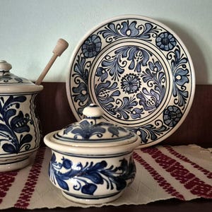 Może przedstawiać: Zestaw ceramiczny składający się z dzbanka na miód z pokrywką i drewnianą łyżeczką, małej cukiernicy z pokrywką oraz ozdobnego talerza. Każdy element jest w kolorze kremowym z ręcznie malowanymi niebieskimi wzorami kwiatów i liści.