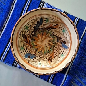 May include: A decorative ceramic bowl with a floral design in shades of orange, brown, and blue. The bowl sits on a blue and white striped textile with fringe. The bowl's rim is a warm brown color.