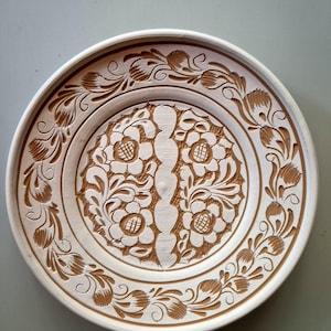 May include: A round, off-white ceramic plate with intricate floral carvings. The design features a central vertical element flanked by symmetrical flower patterns and a border of stylized leaves and blossoms. The plate is a decorative piece.