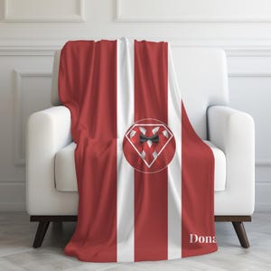 Op de afbeelding: Een rood-wit gestreepte plaid over een witte fauteuil. De deken heeft een ruitvormig embleem met een vlinderdasontwerp op een rode cirkel. Het woord "Dona" staat op de deken.