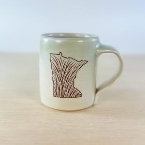 May include: A ceramic mug with a light green gradient. The mug features a brown illustration of the state of Minnesota filled with a textured design. The mug has a curved handle and sits on a light-colored wooden surface.