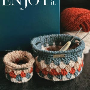 May include: Two handmade crochet baskets in beige, red, and blue. The larger basket has a blue rim and a "Handmade" tag. The smaller basket is similar in design. The image also includes the text "ENJOY it."