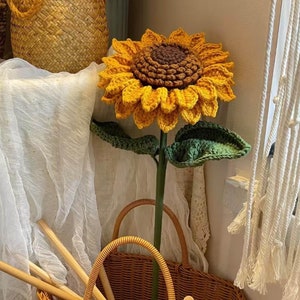 May include: A large, handcrafted crochet sunflower with yellow petals and a brown center. The sunflower has green leaves and a long green stem. A woven basket and other decorative items are in the background.