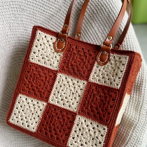May include: A handmade crochet tote bag with a checkerboard pattern in rust and cream colors. The bag features brown leather handles and accents, with metal hardware. The bag is square-shaped and appears to be sturdy and well-crafted.