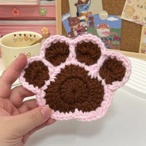 May include: A brown crocheted paw print coaster with a pink border.