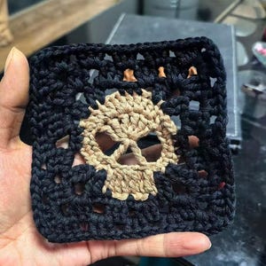 Könnte beinhalten: Ein quadratischer, handgefertigter Häkeluntersetzer mit einem Totenkopf-Design. Der Untersetzer hat einen schwarzen Rand mit einem beigefarbenen Totenkopf in der Mitte. Der Totenkopf ist mit beigem Garn gehäkelt, und der Rand ist schwarz.
