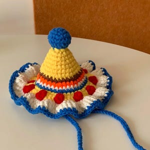 Puede incluir: Un sombrero de ganchillo hecho a mano con un diseño de payaso. El sombrero presenta un cono amarillo con un pompón azul en la parte superior, un borde festoneado blanco y azul y detalles circulares rojos. Lazos azules se extienden desde el borde.