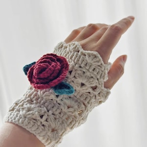 Könnte beinhalten: Cremefarbene Häkel-Fingerhandschuhe mit einer burgunderroten Rose und türkisfarbenen Blättern. Die Handschuhe haben ein strukturiertes Stichmuster und einen gewellten Rand. Der handgefertigte Blumenschmuck verleiht den Handgelenkwärmern eine dekorative Note.