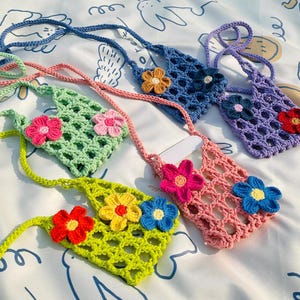 Może przedstawiać: Pięć mini torebek wykonanych na szydełku z kwiatowymi akcentami. Torebki są w różnych kolorach, w tym zielonym, niebieskim, fioletowym i różowym. Każda torebka ma szydełkowy pasek i kwiatowy wzór z przodu.