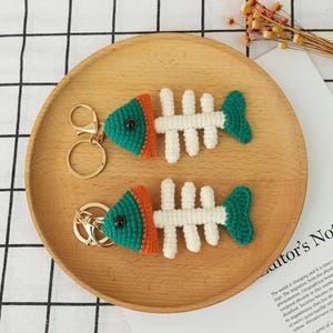 Puede incluir: Dos llaveros de esqueleto de pez de crochet con cuerpos verdes y naranjas y huesos blancos. Los llaveros están en un plato de madera.
