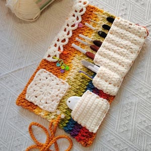 May include: A rainbow-colored crochet organizer roll featuring a textured stitch pattern, a white lace-style border, and attached pockets. It holds several metal crochet hooks, colorful plastic stitch markers, and a small white measuring tool.