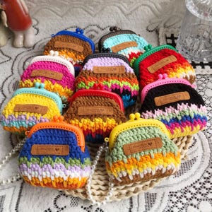 Könnte beinhalten: Eine Sammlung farbenfroher, handgefertigter Häkeltaschen. Jede Tasche hat eine einzigartige Kombination aus leuchtenden Farben, einen Schnappverschluss und ein kleines "Hand Made"-Etikett. Die Taschen sind auf einer dekorativen Oberfläche angeordnet.