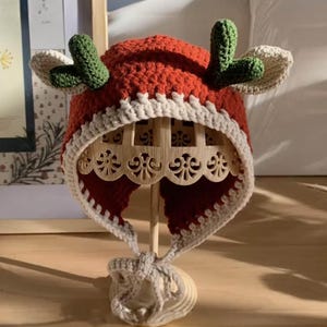 May include: A handmade crochet hat in the shape of a reindeer. The hat is primarily red with a white trim and ear flaps. Green antlers are attached to the top. The hat is displayed on a wooden stand.