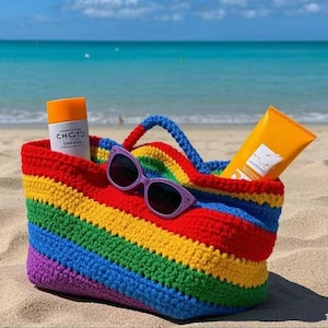 Könnte beinhalten: Eine gehäkelte Tragetasche mit Regenbogenstreifen und blauem Griff, die Sonnenbrille und Sonnencreme enthält. Die Tasche befindet sich an einem Sandstrand mit dem Meer im Hintergrund. Die Sonnenbrille ist lila und die Sonnencreme ist orange.