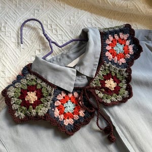 Op de afbeelding: Een kleurrijke gehaakte kraag met een bloemmotief, met tinten rood, blauw, groen en bruin. De kraag wordt getoond op een lichtblauw overhemd, hangend aan een paarse hanger. De kraag heeft een geschulpte rand en bruine strikjes.