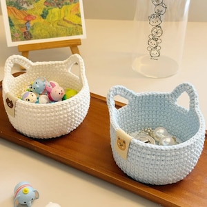Puede incluir: Dos cestas de ganchillo hechas a mano, una blanca y otra azul claro, están sobre una bandeja de madera. Cada cesta tiene asas en forma de oreja y una pequeña etiqueta de oso. La cesta blanca contiene pequeñas figuras, mientras que la cesta azul contiene joyas.