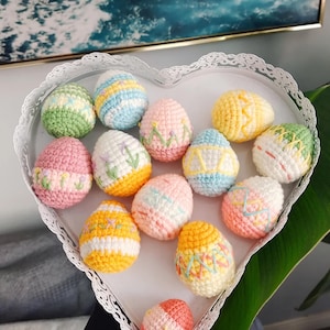 May include: A collection of crocheted Easter eggs in a white heart-shaped dish. The eggs are decorated with various pastel colors and patterns, including pink, blue, yellow, and white.