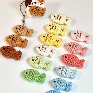 Op de afbeelding: Een verzameling kleurrijke, gehaakte visbedels in verschillende tinten, waaronder bruin, geel, roze, groen en blauw. Elke vis heeft een gedetailleerd schubbenpatroon en een zwart oog. Sommige vissen zijn bevestigd aan een sleutelhanger.
