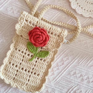 May include: Cream-colored crochet phone bag with a red rose applique and green leaves. The bag has a long strap and a scalloped edge. The crochet pattern features open squares. A white crochet doily is in the background.