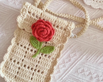 May include: Cream-colored crochet phone bag with a red rose applique and green leaves. The bag has a long strap and a scalloped edge. The crochet pattern features open squares. A white crochet doily is in the background.