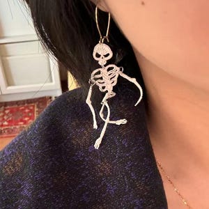 May include: A pair of dangling skeleton earrings. The earrings are white and feature a detailed skeleton design. The earrings hang from gold-colored ear wires. The skeleton's arms and legs are outstretched, giving the impression of movement.