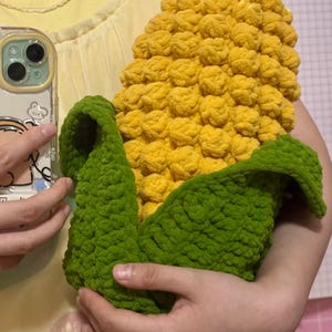 May include: A handmade, plush corn-on-the-cob, with a textured yellow kernel section and green, leafy husks. The soft, crocheted design is held in front of a yellow top.