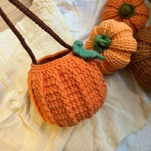 May include: An orange crochet pumpkin-shaped bag with a brown strap and green leaf detail. The bag is part of a collection of handmade crochet pumpkins in various sizes and shades of orange.